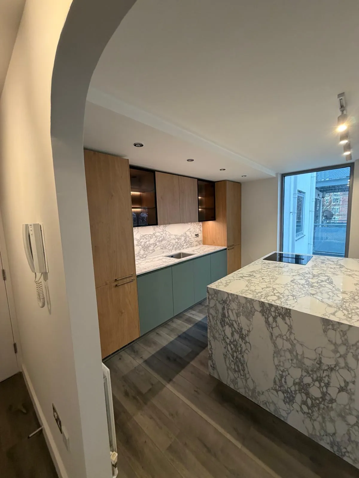 A finished kitchen: a marble-topped island, sage-green and oak units, and a tall window onto the street